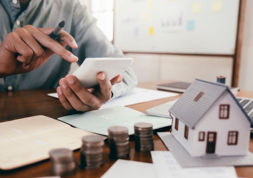 person using a calculator with a stack of coins and house figurine in front of them